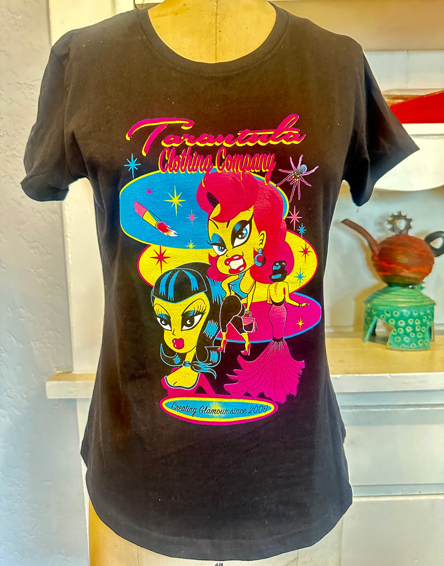 Limited edition women’s cut tee featuring Tarantula Clothing Company Glamour Dolls graphic with colorful retro characters and bold anniversary artwork.